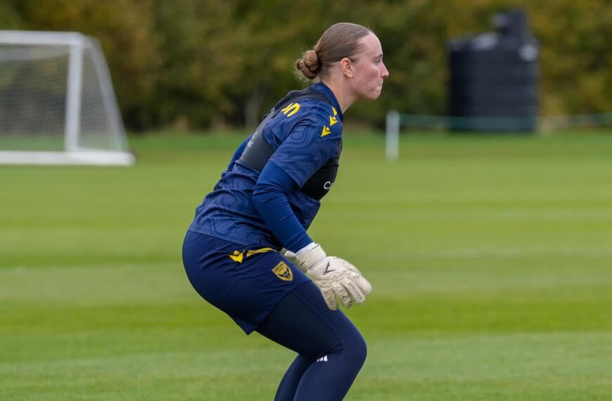 A side profile of Maddison Millington-Stanbury, goalkeeper, during a training session. The player is in a focused, crouched "ready position" on a green grass field. She is wearing a navy blue long-sleeved training top, navy leggings, white goalkeeper gloves, and white boots. Her hair is pulled back into a neat bun. In the background, there is a white goal and a line of green and yellow trees under an overcast sky.