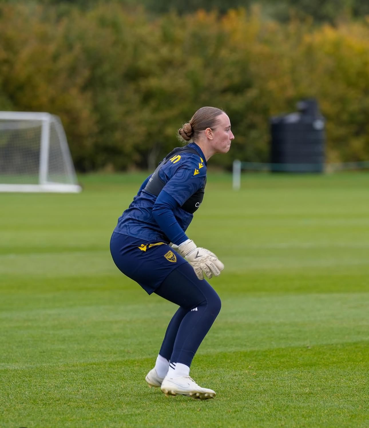 A side profile of Maddison Millington-Stanbury, goalkeeper, during a training session. The player is in a focused, crouched "ready position" on a green grass field. She is wearing a navy blue long-sleeved training top, navy leggings, white goalkeeper gloves, and white boots. Her hair is pulled back into a neat bun. In the background, there is a white goal and a line of green and yellow trees under an overcast sky.
