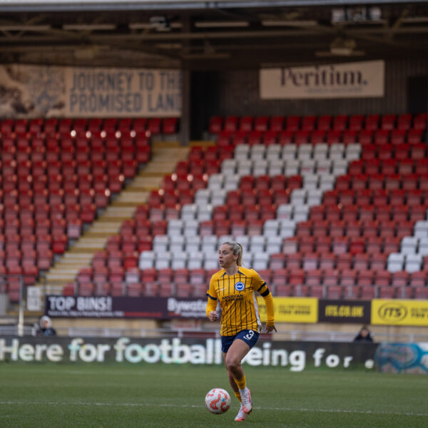 Poppy Pattinson with her hair in a ponytail, wearing a yellow and blue striped kit and blue shorts, dribbles a ball across a green field. She is positioned in the centre of the frame against a backdrop of a large, mostly empty stadium with red and white seats. Advertisements and banners are visible on the stadium walls behind her.
