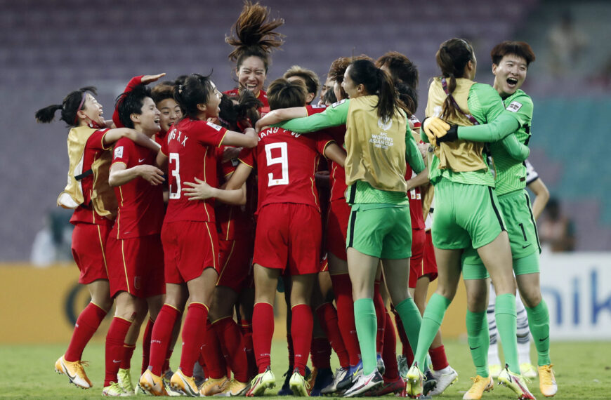 China's Women’s national team players in red kits celebrating together in a large huddle on the pitch, smiling and embracing while two goalkeepers in green hug on the side during the post-match celebration.