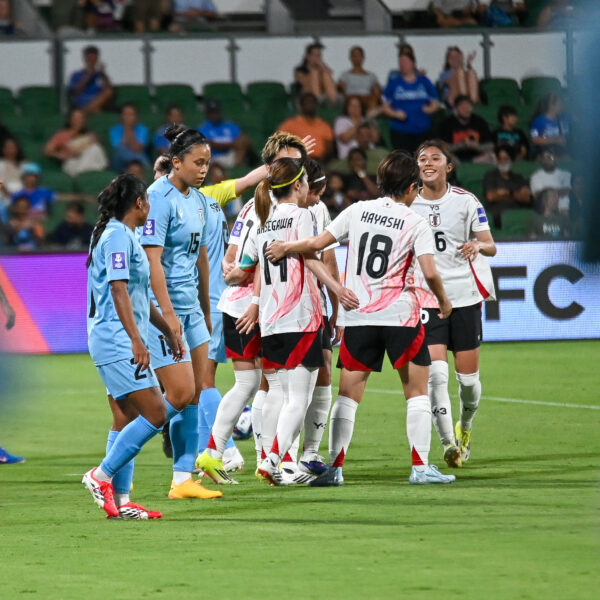 A wide shot from a low angle on a football field shows a group of female players from two teams. In the centre, a group of players in white kits with pink abstract patterns and black shorts are huddling and celebrating. One player, wearing kit number 18 with the name "HAYASHI" on the back, and another with number 6 are visible. To the left, several players in light blue jerseys look on with neutral or disappointed expressions. The game is taking place at night in a stadium with green seating and spectators in the background. The foreground is slightly blurred with the shapes of people out of focus.