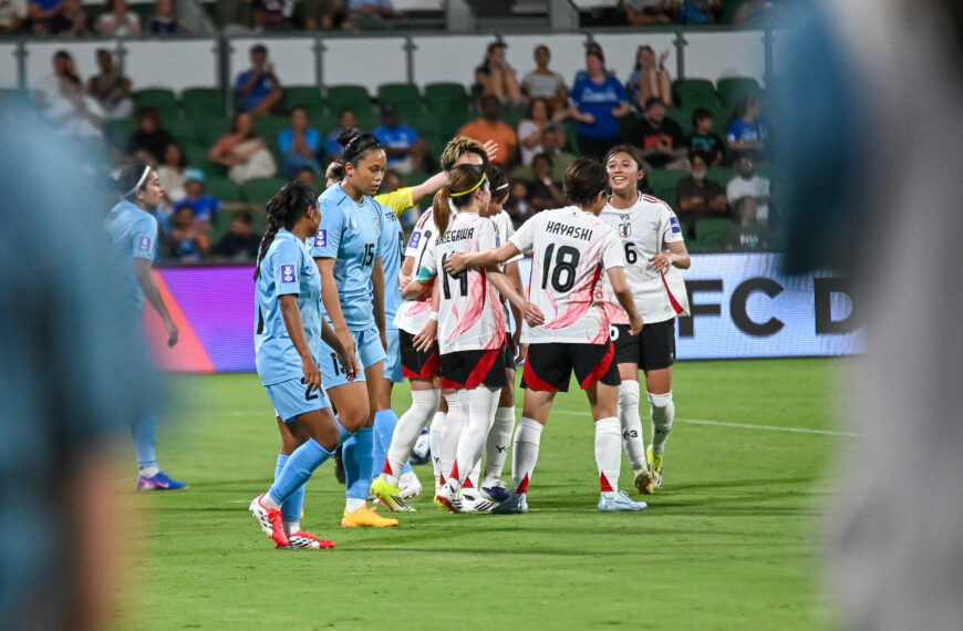 A wide shot from a low angle on a football field shows a group of female players from two teams. In the centre, a group of players in white kits with pink abstract patterns and black shorts are huddling and celebrating. One player, wearing kit number 18 with the name "HAYASHI" on the back, and another with number 6 are visible. To the left, several players in light blue jerseys look on with neutral or disappointed expressions. The game is taking place at night in a stadium with green seating and spectators in the background. The foreground is slightly blurred with the shapes of people out of focus.