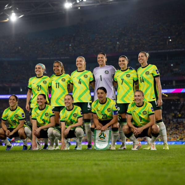 Eleven players of the Australian women’s national team (the Matildas) pose for a group photo on a brightly lit grass pitch at night. Six players stand in the back row, and five kneel in the front row. They are wearing their vibrant yellow and green patterned home kits with dark green shorts, except for the goalkeeper in the centre of the back row, who is wearing a light purple jersey. The captain, Sam Kerr (number 20), kneels in the centre of the front row holding a team pennant. A massive, blurred crowd fills the stands of the stadium under bright stadium lights in the background.