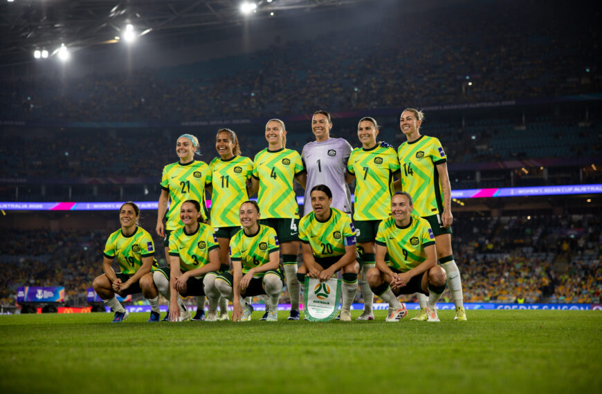 Eleven players of the Australian women’s national team (the Matildas) pose for a group photo on a brightly lit grass pitch at night. Six players stand in the back row, and five kneel in the front row. They are wearing their vibrant yellow and green patterned home kits with dark green shorts, except for the goalkeeper in the centre of the back row, who is wearing a light purple jersey. The captain, Sam Kerr (number 20), kneels in the centre of the front row holding a team pennant. A massive, blurred crowd fills the stands of the stadium under bright stadium lights in the background.