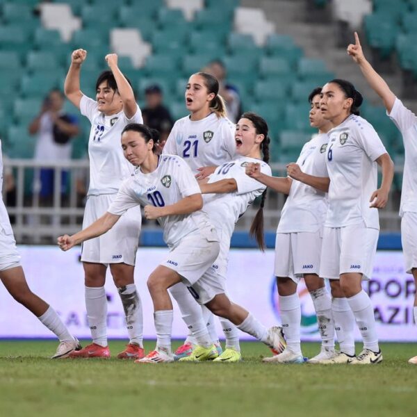 An action-oriented image capturing a moment of intense celebration by a women's football team. The players, wearing matching white uniforms with blue numbering and the "UFA" (Uzbekistan Football Association) crest, are spread across a green pitch in a stadium. Their expressions and gestures convey pure triumph: some have their fists clenched and raised high, others are shouting in joy, and one player on the far right points upward with both hands. The stadium seating in the background is a mix of green and white, though blurred, keeping the focus entirely on the emotional energy of the athletes on the field.