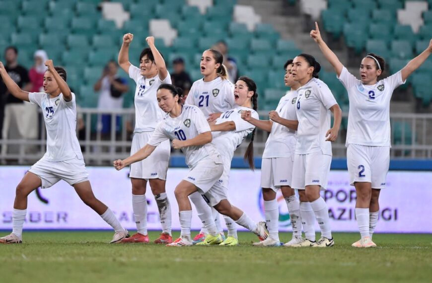 An action-oriented image capturing a moment of intense celebration by a women's football team. The players, wearing matching white uniforms with blue numbering and the "UFA" (Uzbekistan Football Association) crest, are spread across a green pitch in a stadium. Their expressions and gestures convey pure triumph: some have their fists clenched and raised high, others are shouting in joy, and one player on the far right points upward with both hands. The stadium seating in the background is a mix of green and white, though blurred, keeping the focus entirely on the emotional energy of the athletes on the field.