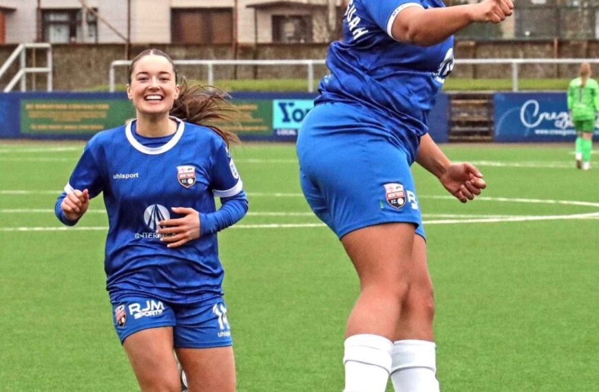 A high-action, medium shot of two female players from Montrose F.C. celebrating on a bright green turf field. One player on the right is captured mid-air in a dynamic jump, her body angled sideways with a joyful, shouting expression and a clenched fist. Behind her, another player runs toward her with a wide, beaming smile. Both are wearing matching blue kits with white trim and white socks. The background shows a suburban setting with houses and a perimeter fence under an overcast sky.