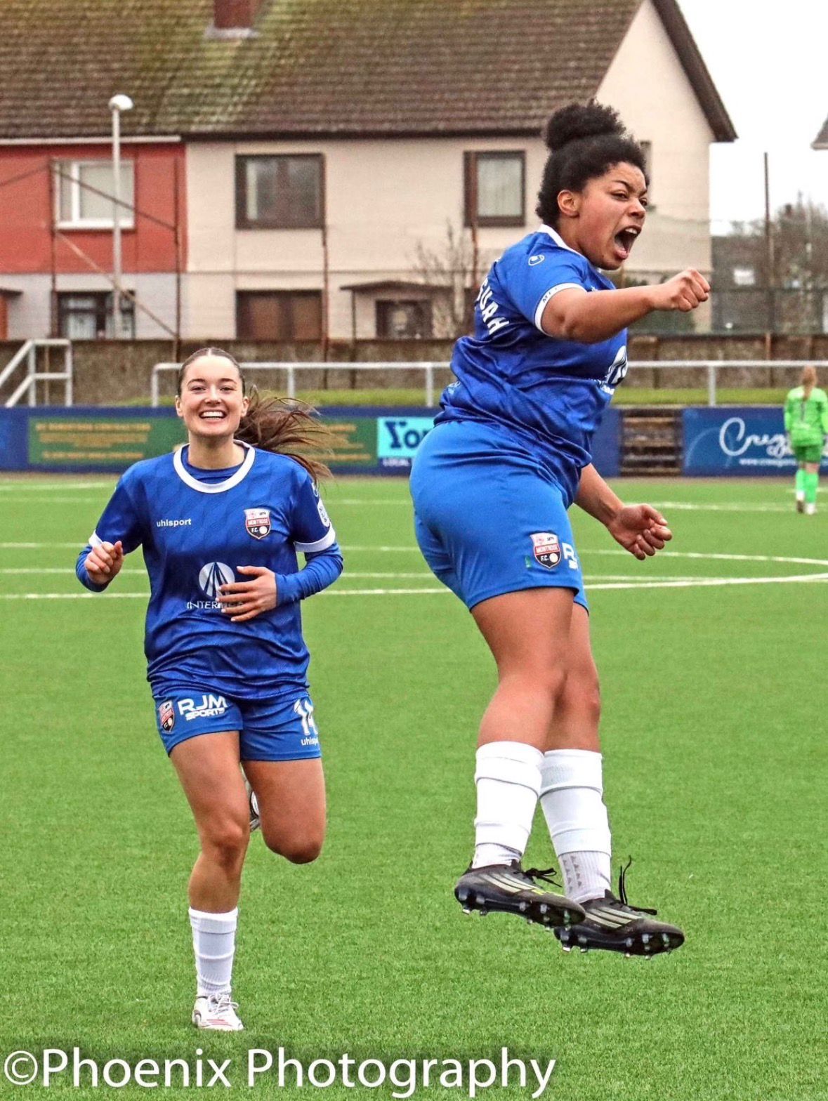 A high-action, medium shot of two female players from Montrose F.C. celebrating on a bright green turf field. One player on the right is captured mid-air in a dynamic jump, her body angled sideways with a joyful, shouting expression and a clenched fist. Behind her, another player runs toward her with a wide, beaming smile. Both are wearing matching blue kits with white trim and white socks. The background shows a suburban setting with houses and a perimeter fence under an overcast sky.