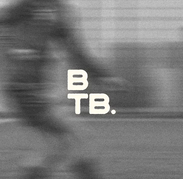 A grainy, black-and-white action photograph with a heavy horizontal motion blur, suggesting high speed. The blurred silhouette of an athlete appears to be moving across a field or track. In the centre, the letters "B" and "TB." are stacked vertically in a bold, white, slightly distressed sans-serif font.