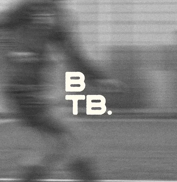A grainy, black-and-white action photograph with a heavy horizontal motion blur, suggesting high speed. The blurred silhouette of an athlete appears to be moving across a field or track. In the centre, the letters "B" and "TB." are stacked vertically in a bold, white, slightly distressed sans-serif font.