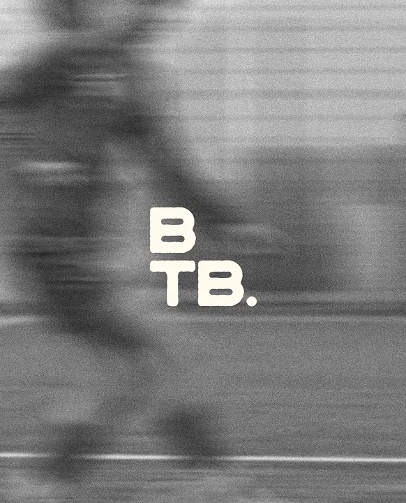 A grainy, black-and-white action photograph with a heavy horizontal motion blur, suggesting high speed. The blurred silhouette of an athlete appears to be moving across a field or track. In the centre, the letters "B" and "TB." are stacked vertically in a bold, white, slightly distressed sans-serif font.