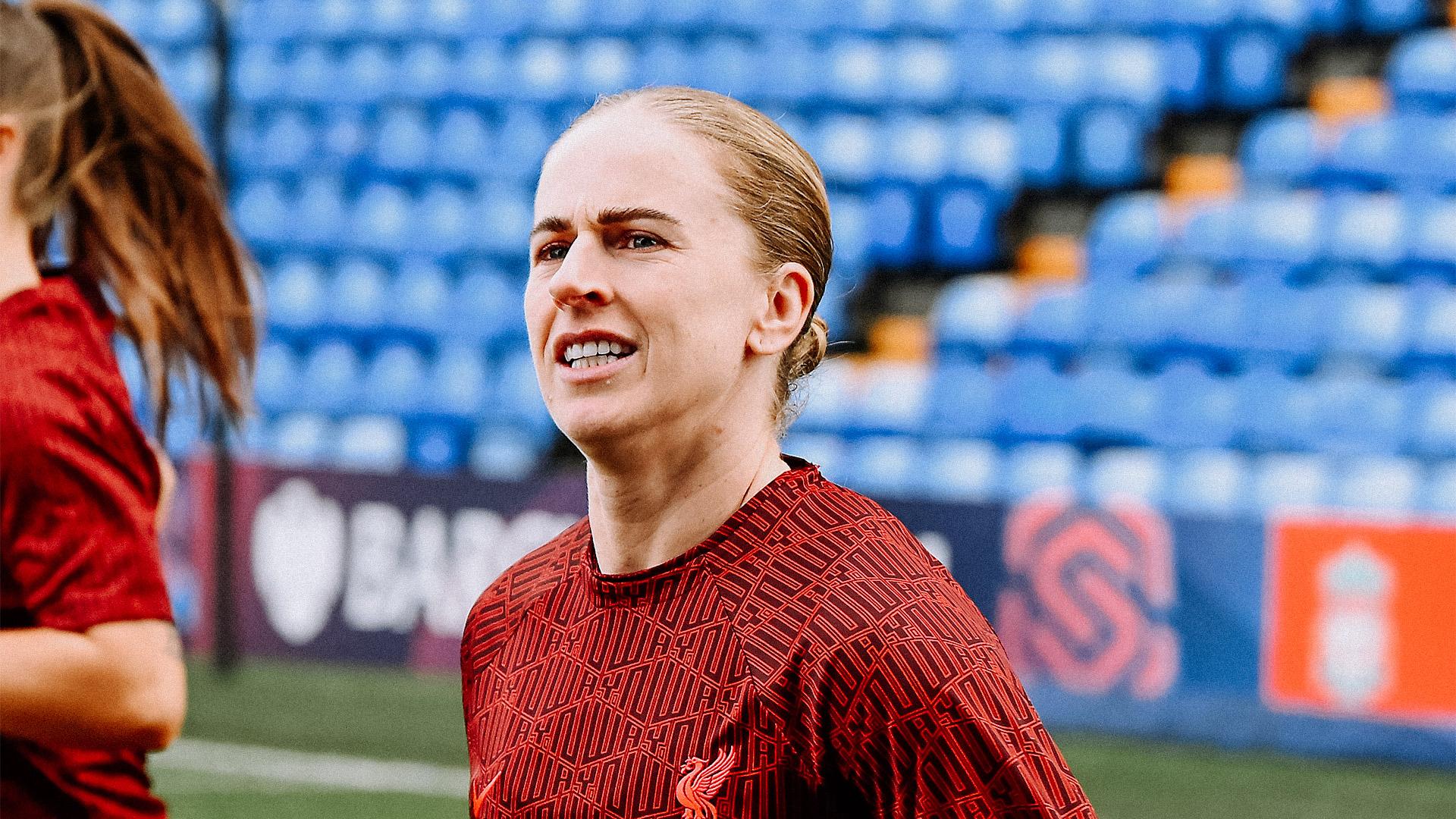 A close-up shot of professional football player Natasha Dowie on a pitch. She is wearing a maroon Liverpool FC training top featuring a geometric pattern and the club's Liver Bird crest. Her blonde hair is pulled back into a bun, and she has a focused expression while looking off-camera. The background shows blurred blue and orange stadium seating.
