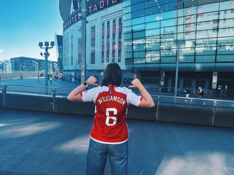 A person stands with their back to the camera outside the Emirates Stadium, pointing both thumbs towards the name "Williamson" and number 6 on the back of an Arsenal home shirt. The stadium's glass exterior and entrance dominate the background, with a few people walking in the distance on a bright, clear day. The person is wearing blue jeans and stands on a raised walkway overlooking the stadium forecourt.