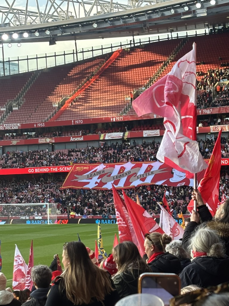 Supporters in the stands at Emirates Stadium wave red, white, and pink Arsenal flags before a match. A large banner reading “Leader. Legend. Little.” is displayed across the crowd in tribute to Kim Little, while sunlight streams through the stadium roof onto the opposite stand. The pitch is visible in the foreground, creating a vibrant matchday atmosphere.