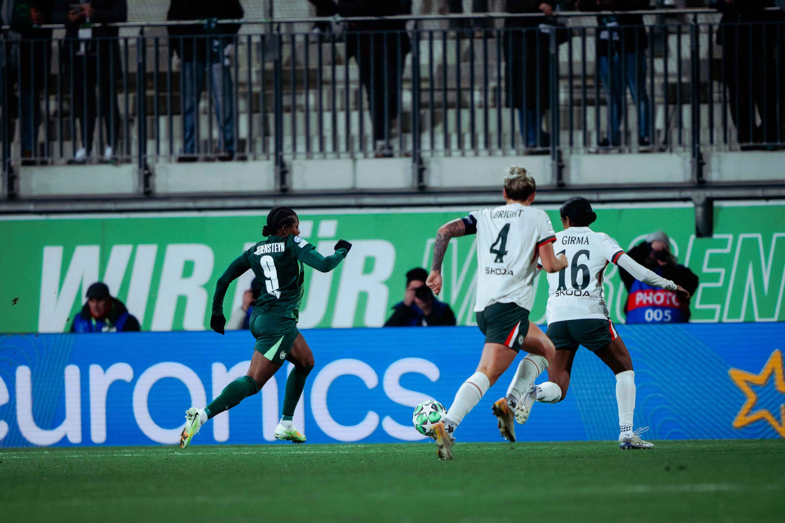 Lineth Beerensteyn runs forward in a green Wolfsburg kit during a UEFA Women’s Champions League match, pursued by Chelsea defenders Millie Bright and Naomi Girma.