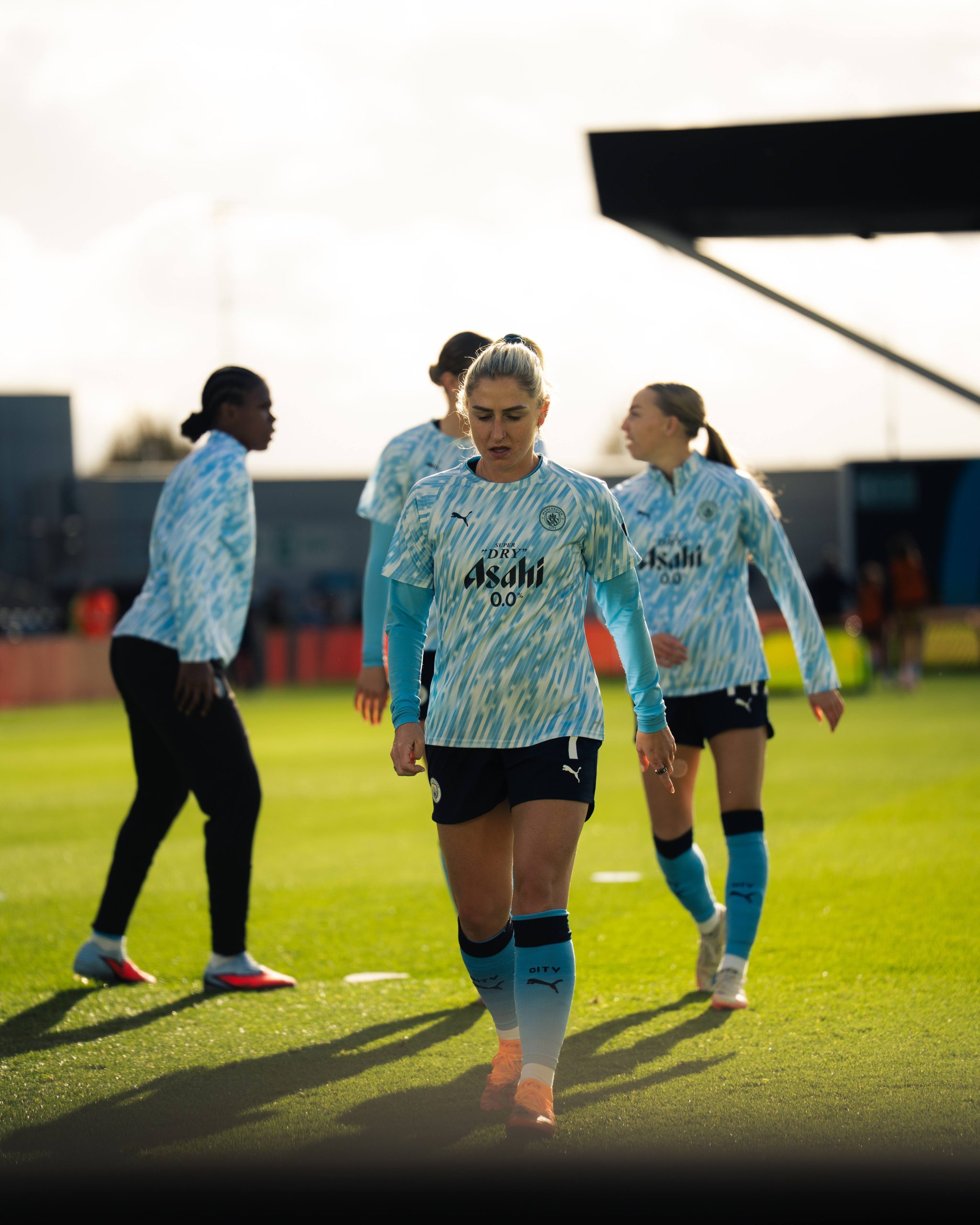 Manchester City Women players warm up on a sunlit training pitch before a match. One player in the foreground, Laura Coombs, wearing Manchester City’s light blue patterned warm-up top and navy shorts, walks across the grass with her head slightly down, while teammates move and stretch in the background. The low evening sunlight creates long shadows across the pitch and gives the scene a warm, focused pre-match atmosphere.