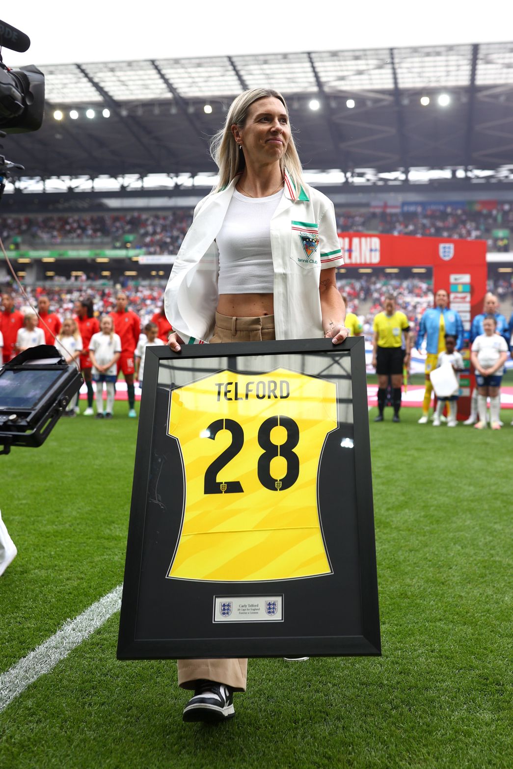 Former England goalkeeper Carly Telford walks onto the pitch at a packed stadium while holding a framed yellow England shirt bearing her name and number 28. Standing on the touchline before kick-off, she smiles as players, mascots and match officials line up in the background. Stadium lights shine overhead, creating a celebratory atmosphere during a special presentation in recognition of her contribution to English football.