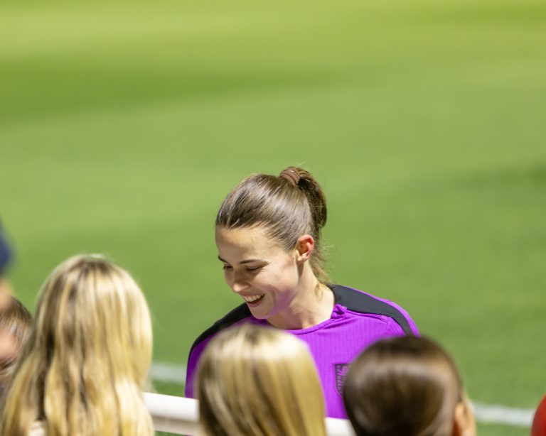 Anouk Denton, footballer, wearing a purple England training top smiles and chats with a group of young supporters beside the pitch. Her hair is tied back in a bun as she leans towards the fans, who are gathered at the barrier. The blurred green playing surface fills the background, creating a warm and friendly post-training atmosphere.