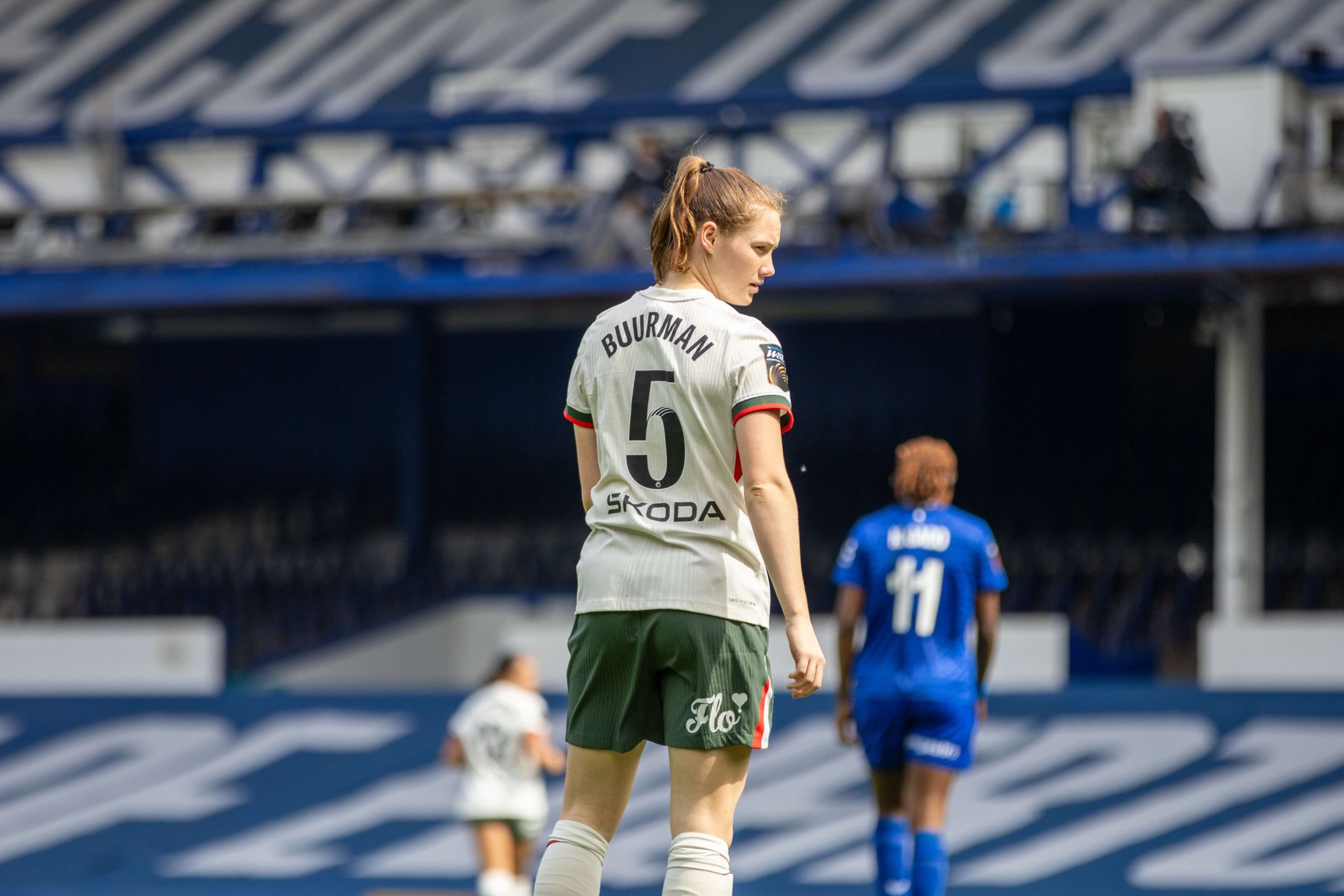 A Chelsea Women player Veerle Buurman wearing the number 5 shirt stands on the pitch during a match, looking across the field. Photographed from behind, she is in sharp focus against a blurred stadium backdrop, with opposition players visible in the distance. The image captures a moment of concentration during play in bright daylight.