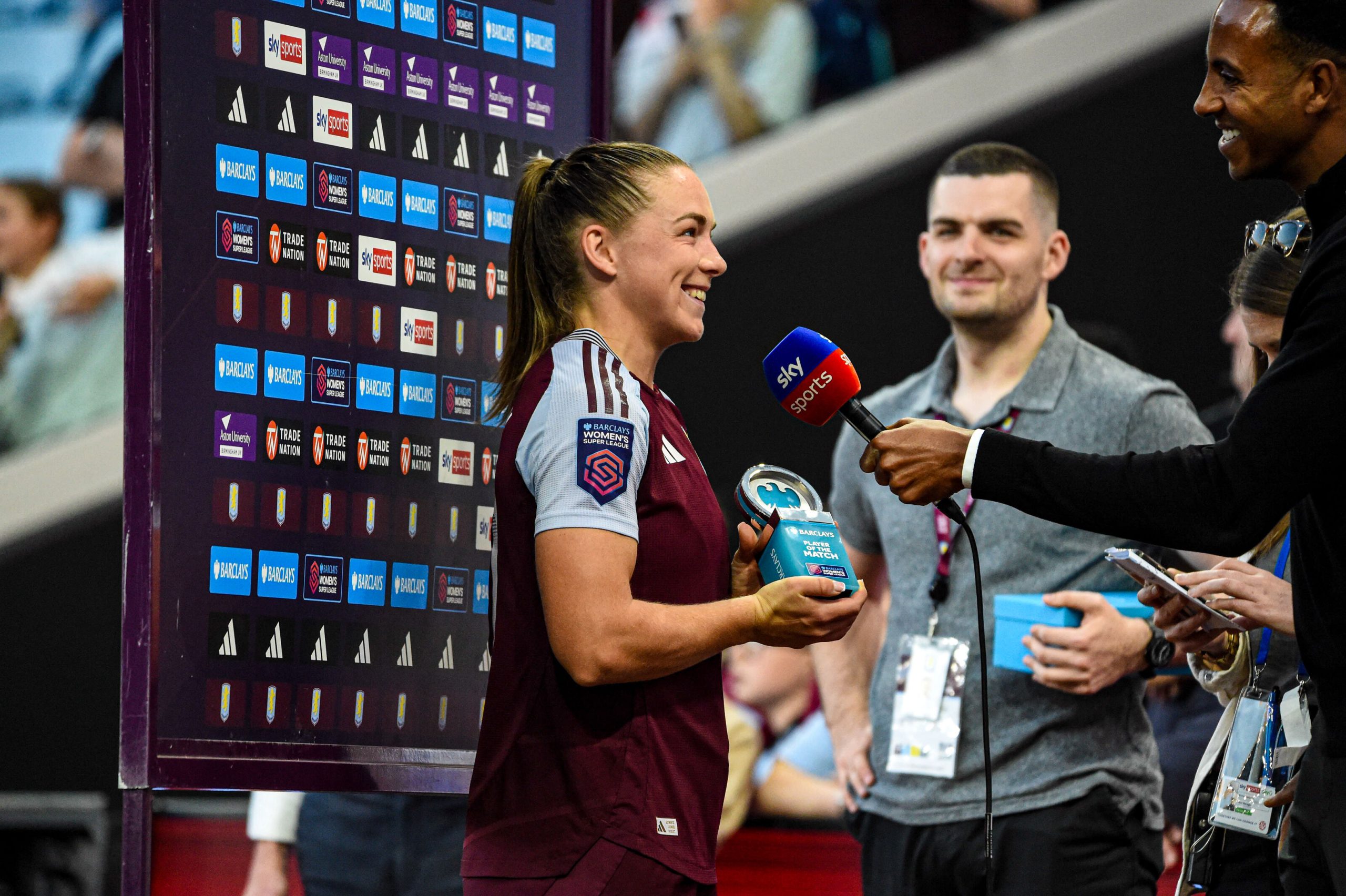 Kirsty Hanson smiles during a post-match interview after receiving the Barclays Women's Super League Player of the Match award for Aston Villa. Holding the trophy, she speaks into a Sky Sports microphone in front of the league's branded interview backdrop while media staff look on at the stadium.