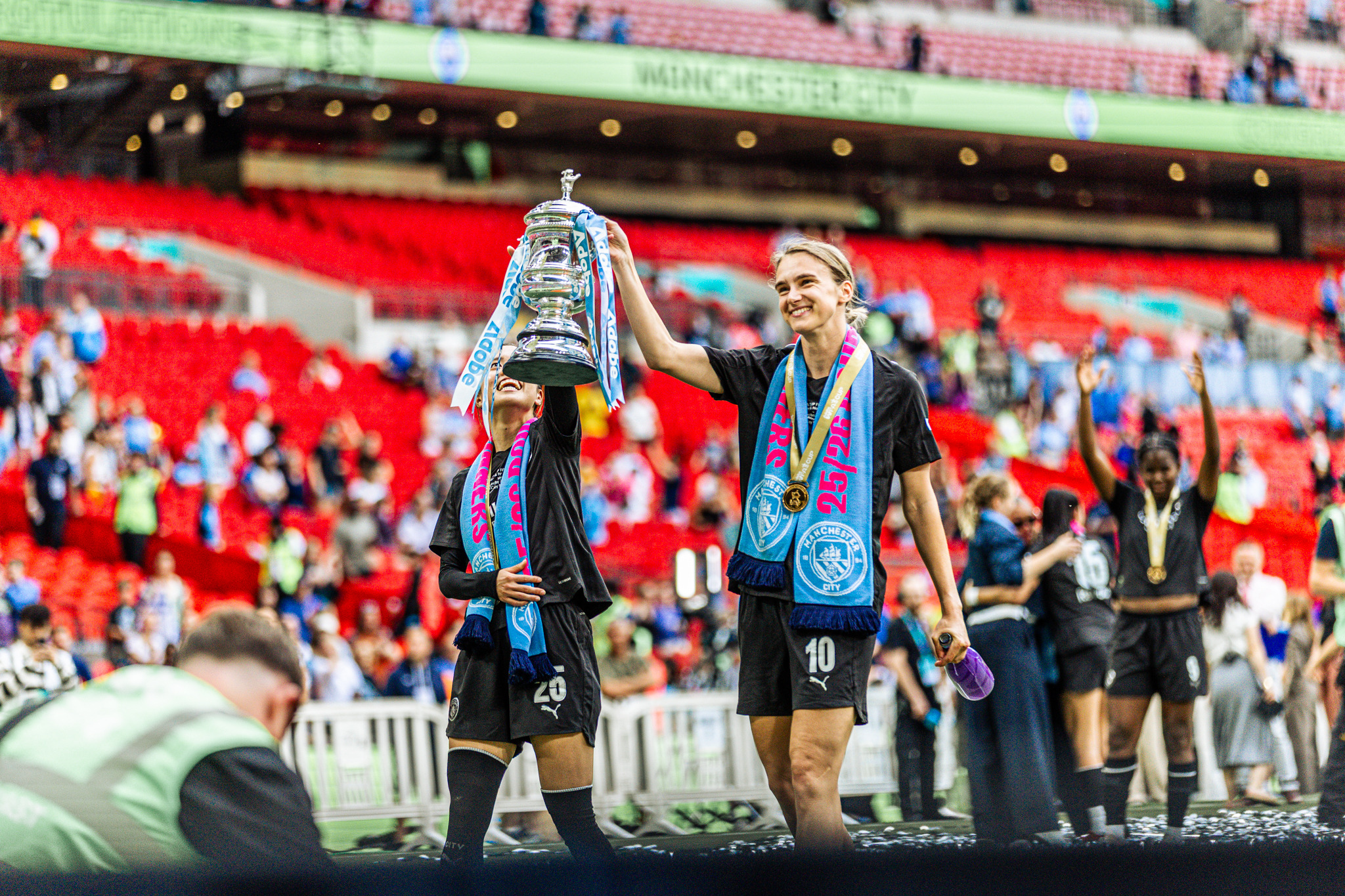 Manchester City players celebrate on the Wembley pitch after winning the 2026 Adobe Women’s FA Cup. One player lifts the trophy aloft while Vivianne Miedema, wearing a Manchester City scarf and winners’ medal, smiles beside her. Teammates celebrate in the background with the red Wembley seats filling the scene.