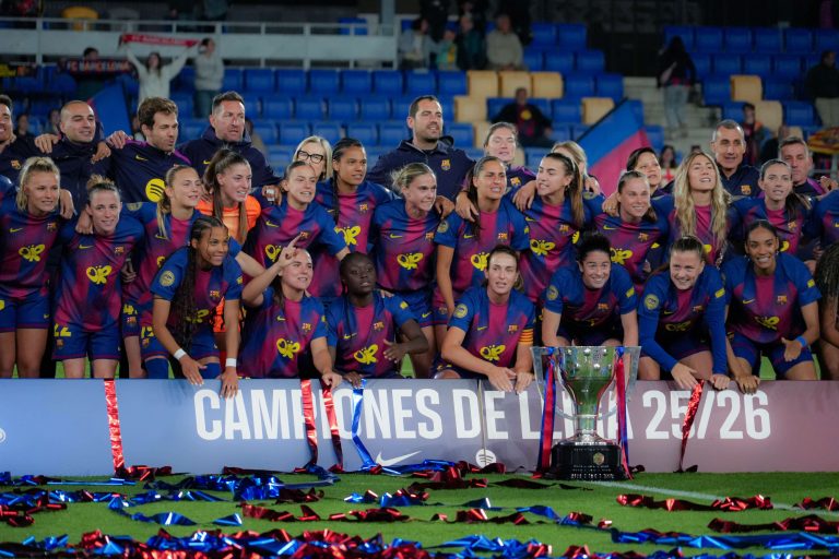 Barcelona Femení players and coaching staff pose together for a team photo after winning the Liga F title. Gathered behind the league trophy on the pitch, the squad wear Barcelona’s blue and maroon kit as red and blue confetti is scattered across the grass. A banner reading “Campeones de Liga 25/26” stands in front of the group, while supporters watch from the stadium stands in the background.