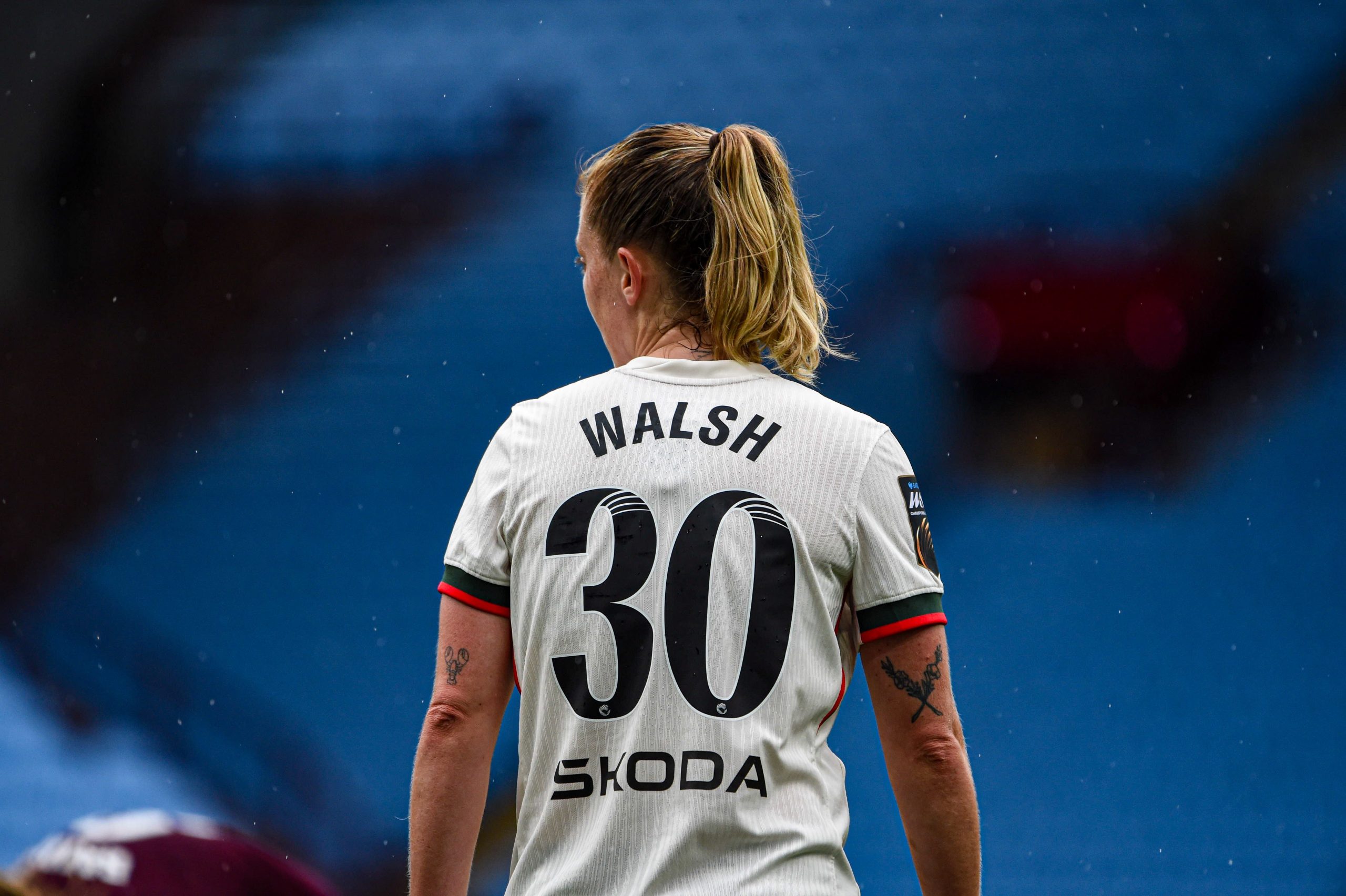 Back view of England midfielder Keira Walsh wearing the number 30 shirt during a match. Walsh stands on the pitch with her blonde hair tied in a ponytail, while light rain falls around her. The white England kit contrasts against the blurred blue stadium seats in the background, creating a focused and atmospheric image.