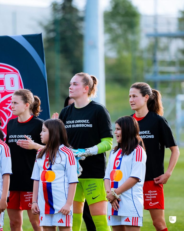 Players from Dijon Women stand alongside child mascots before a match. Several players wear black protest T-shirts with the French slogan “Vous dirigez, nous subissons” (“You lead, we suffer”), while the goalkeeper stands out in a bright green kit and gloves. The mascots wear white shirts with a red and blue vertical stripe, and the group gathers on the pitch during the pre-match ceremony, highlighting the team’s public protest against the club’s treatment of the women’s side.