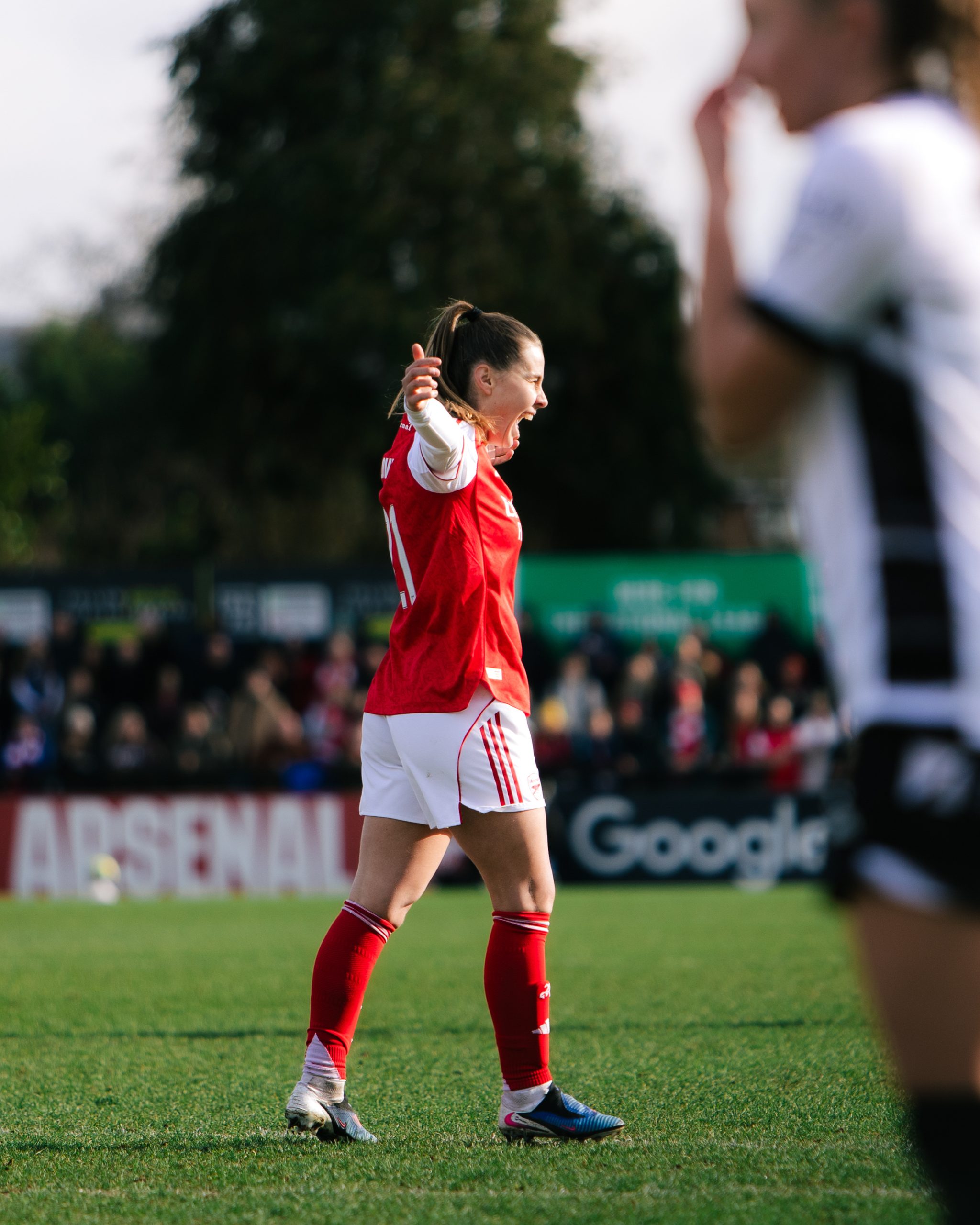 Victoria Pelova of Arsenal celebrates passionately during a match, standing on the pitch with her mouth open in a shout and both arms outstretched. Wearing Arsenal's red and white home kit, she is sharply in focus at the centre of the image, while an opposition player in a black and white kit appears blurred in the foreground and spectators can be seen out of focus in the background.