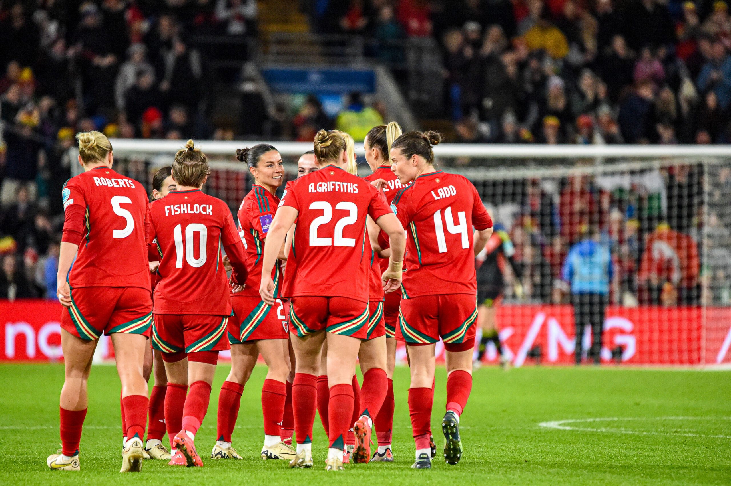 Several Wales women's national team players in red kits gather together in celebration on the pitch, viewed mostly from behind. Players including Angharad James, Sophie Ingle, Carrie Jones and Lily Woodham are visible among the group, with a packed crowd and the goal in the background under the floodlights.