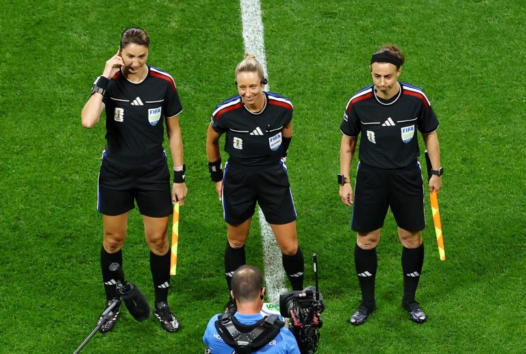 Three female match officials stand on the halfway line of a football pitch before kick-off. The referee in the centre smiles with her hands behind her back, while the two assistant referees stand either side holding brightly coloured flags. All three wear black FIFA referee kits with communication headsets. In the foreground, a television camera operator and pitchside microphone can be seen filming the officials.