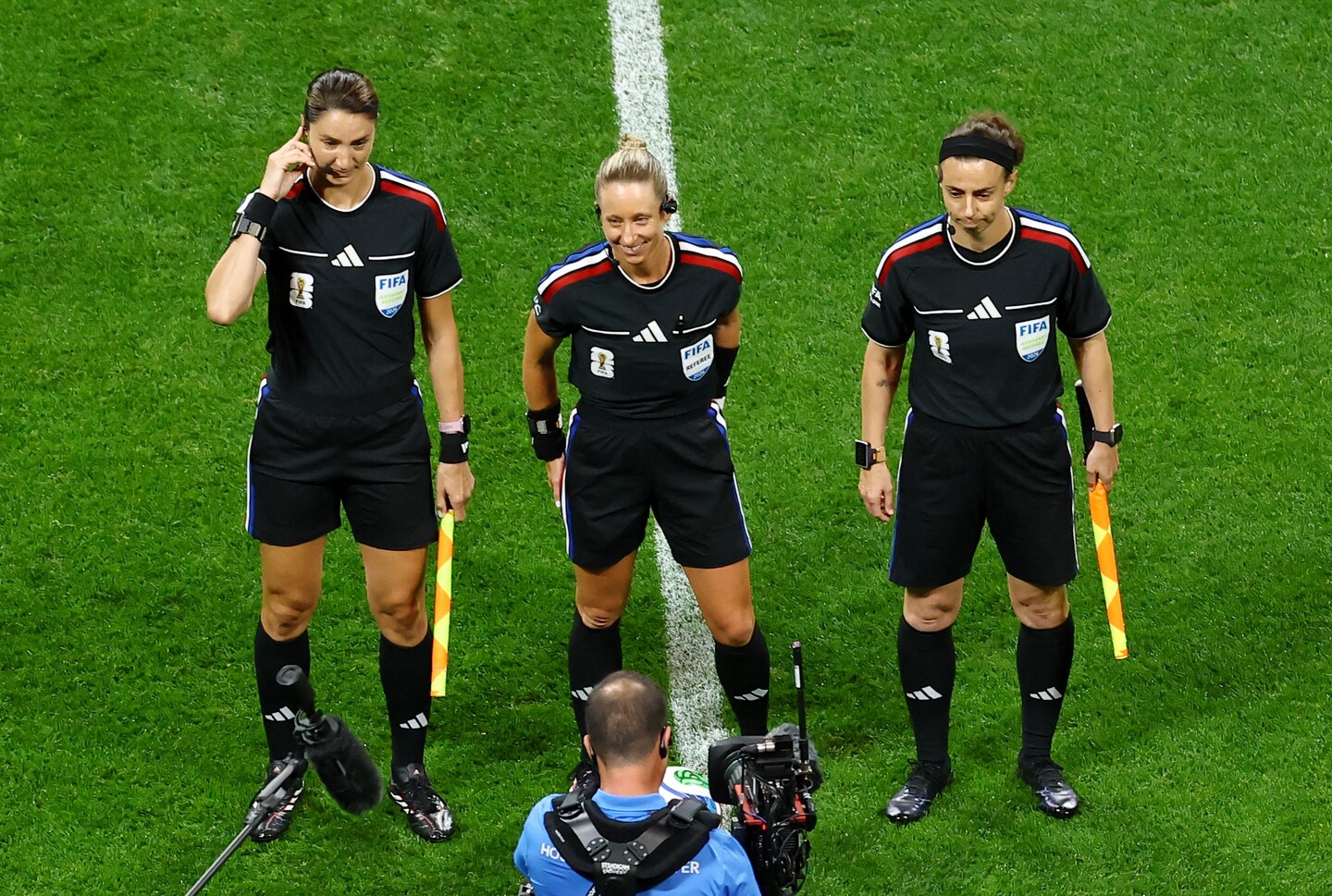 Three female match officials stand on the halfway line of a football pitch before kick-off. The referee in the centre smiles with her hands behind her back, while the two assistant referees stand either side holding brightly coloured flags. All three wear black FIFA referee kits with communication headsets. In the foreground, a television camera operator and pitchside microphone can be seen filming the officials.