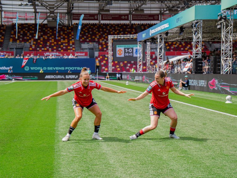 Two Manchester United players in red kits recreate the Macarena dance on the pitch during the World Sevens Football tournament at Brentford's Gtech Community Stadium. The pair mirror each other's movements with arms outstretched, while World Sevens branding, spectators and a scoreboard showing Manchester United leading Everton 1-0 are visible in the background.