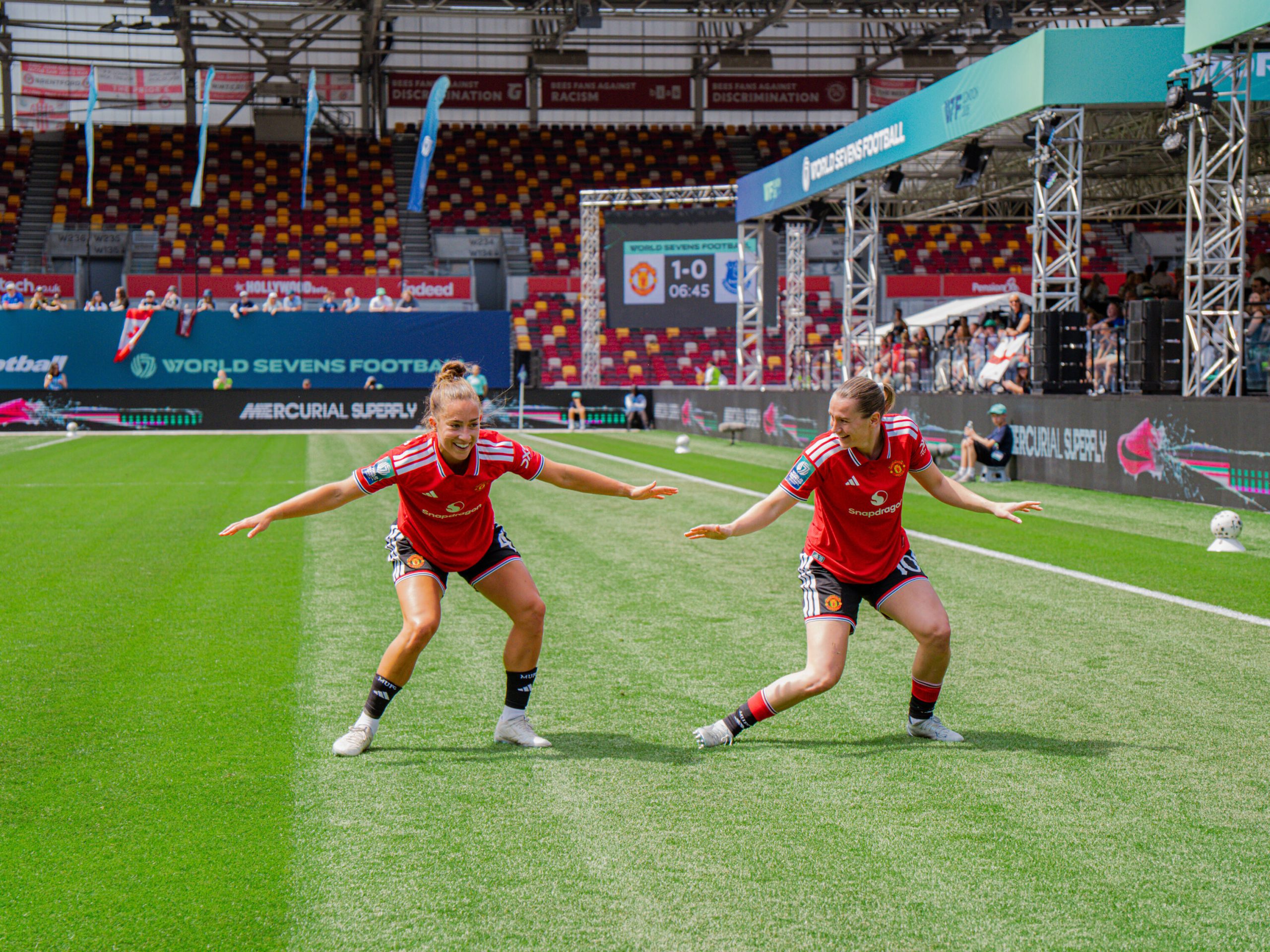 Two Manchester United players in red kits recreate the Macarena dance on the pitch during the World Sevens Football tournament at Brentford's Gtech Community Stadium. The pair mirror each other's movements with arms outstretched, while World Sevens branding, spectators and a scoreboard showing Manchester United leading Everton 1-0 are visible in the background.