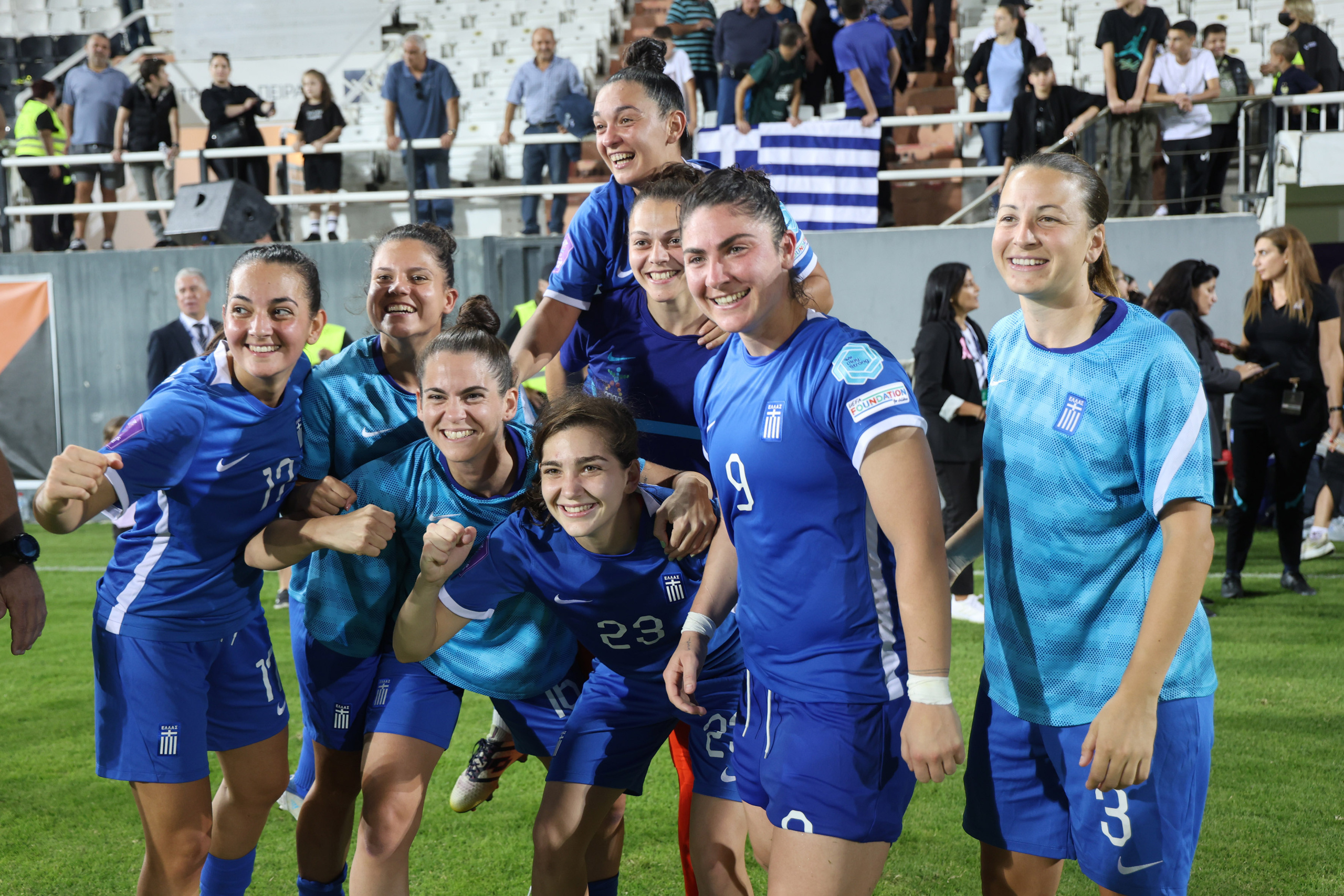A group of eight Greece women’s national team players in blue kits pose together on the pitch after a match, smiling and celebrating for a photo. Several players raise clenched fists in celebration while teammates lean on each other. In the background, supporters stand in the stadium, including fans holding a Greek flag.