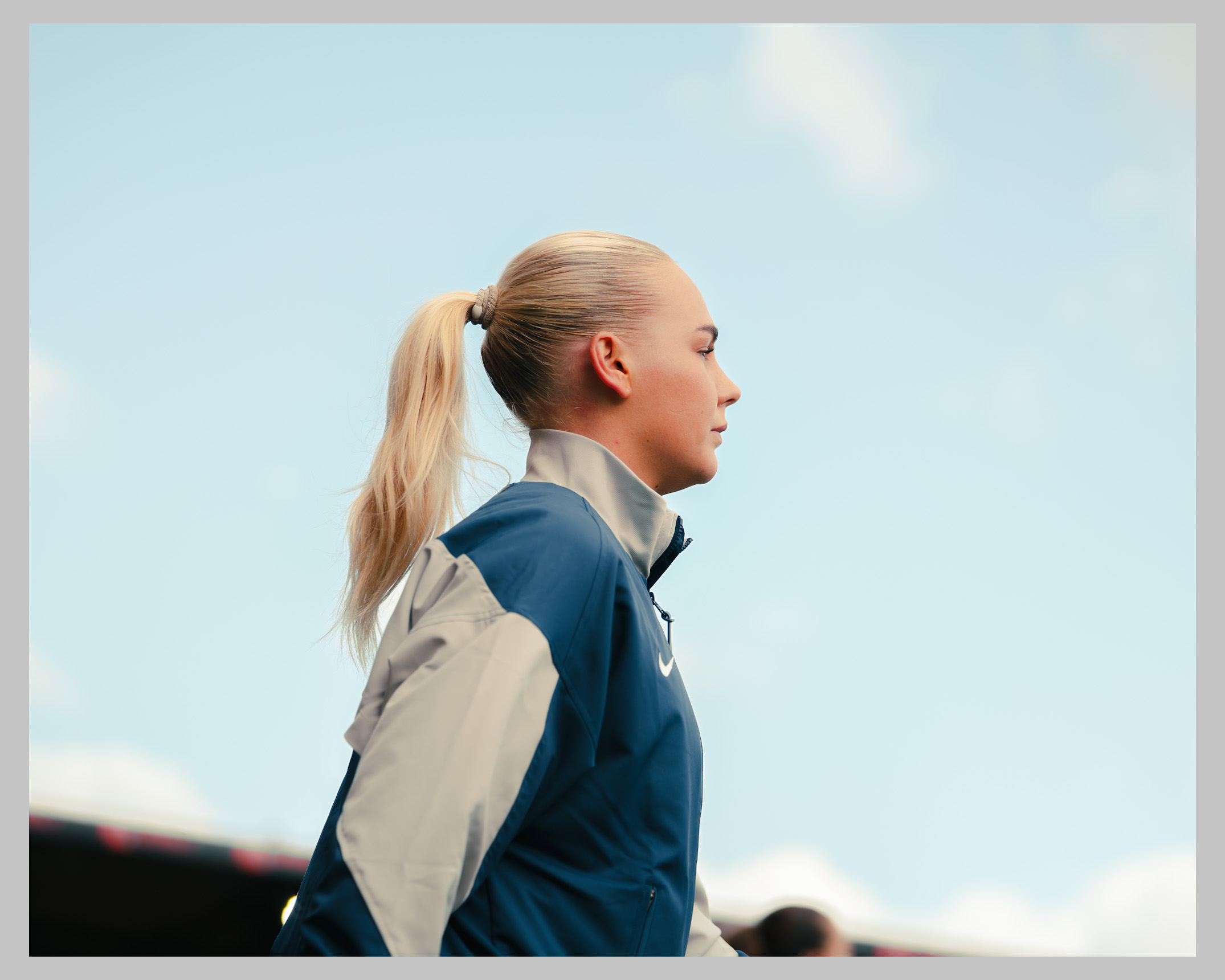 Side-profile portrait of Tottenham Hotspur Women forward Cathinka Tandberg wearing a navy and grey training jacket, with her blonde hair tied back in a ponytail, looking ahead against a clear blue sky before a match.