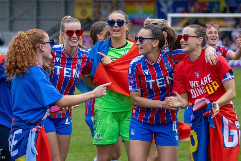 Crystal Palace Women players celebrate promotion together on the pitch, smiling and laughing while wearing heart-shaped sunglasses and waving flags. Several players are dressed in the club's red and blue home kit, while one wears a red "We Are Going Up" T-shirt and another is in a green goalkeeper kit. The joyful group embraces and points at one another as supporters and teammates celebrate in the background.