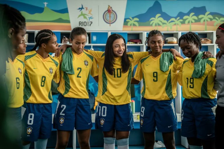Brazil players (actors) stand arm-in-arm in a locker room before a match, wearing yellow shirts and blue shorts as they gather in a team huddle. A player wearing the number 10 shirt smiles while addressing her teammates, with Rio 2016-inspired artwork visible above the lockers in the background.