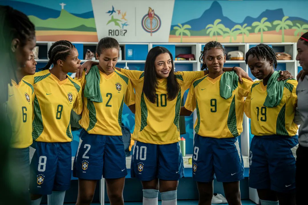 Brazil players (actors) stand arm-in-arm in a locker room before a match, wearing yellow shirts and blue shorts as they gather in a team huddle. A player wearing the number 10 shirt smiles while addressing her teammates, with Rio 2016-inspired artwork visible above the lockers in the background.