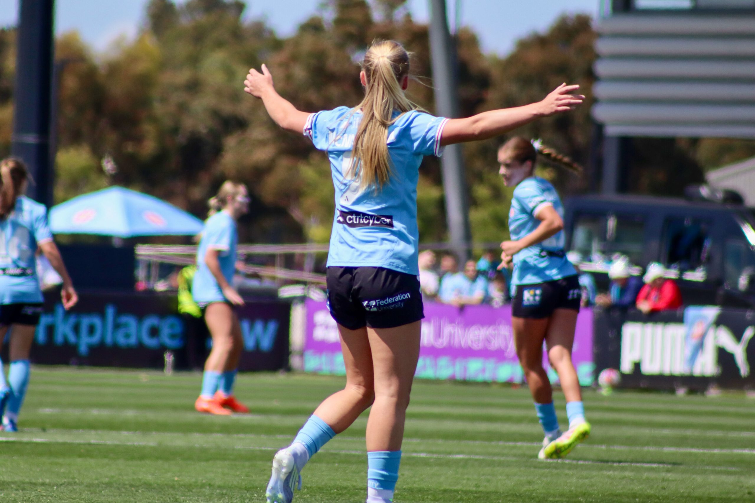 Holly McNamara in a light blue kit stands with her arms outstretched, directing teammates during a match. Viewed from behind, she is positioned in the centre of the pitch while other players move into position in the background on a bright, sunny day.