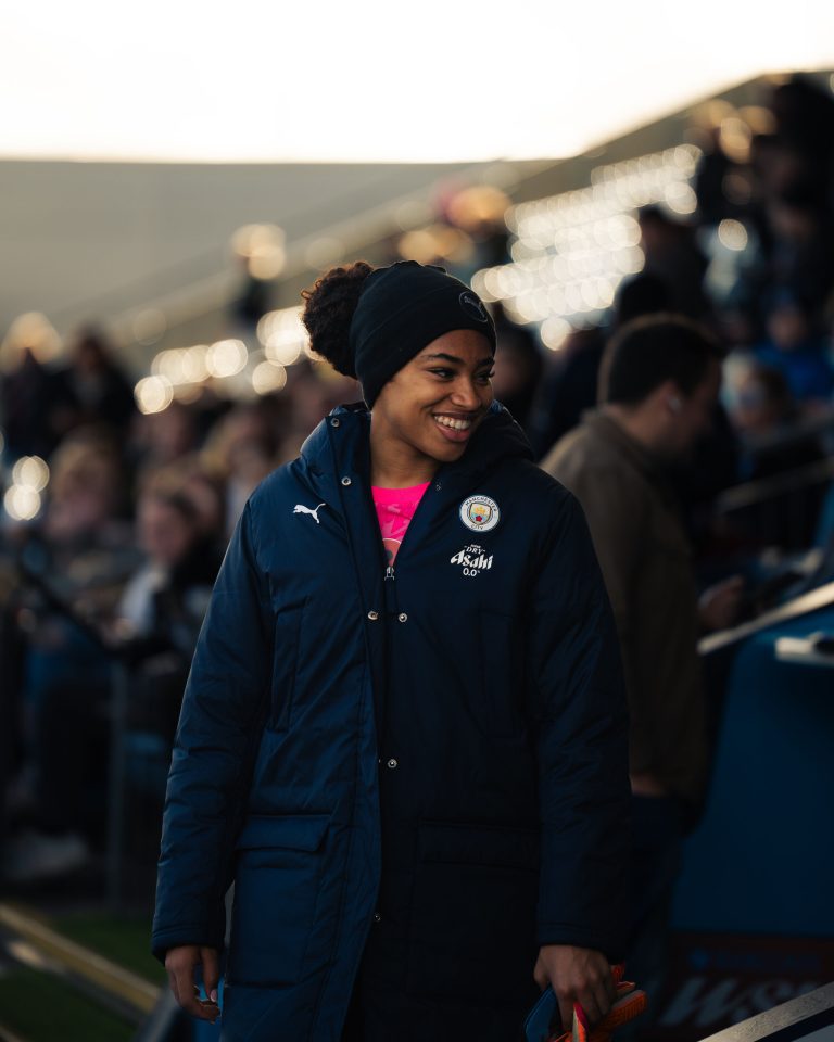 Khiara Keating smiles as she walks along the touchline before a match, wearing a navy Manchester City winter coat and black beanie over a bright pink goalkeeper kit. The blurred stadium crowd and glowing lights in the background create a warm pre-match atmosphere.