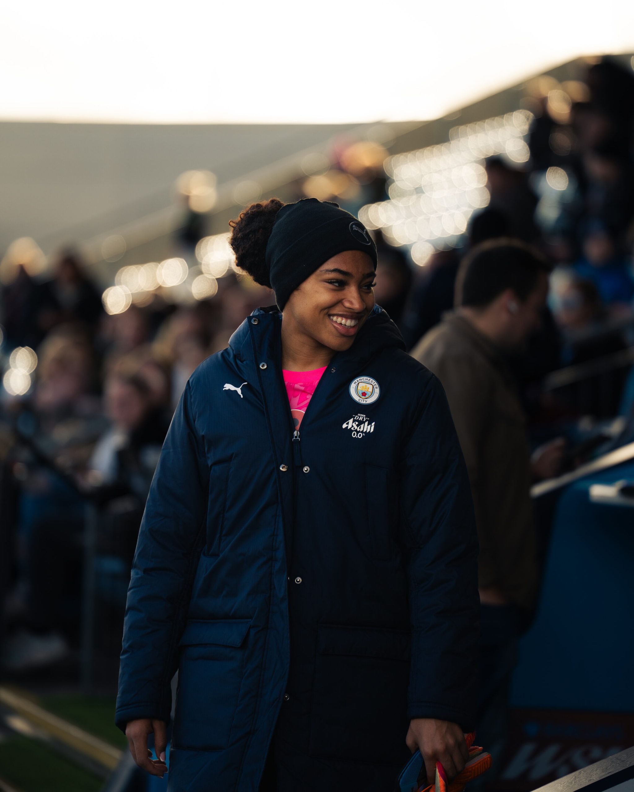 Khiara Keating smiles as she walks along the touchline before a match, wearing a navy Manchester City winter coat and black beanie over a bright pink goalkeeper kit. The blurred stadium crowd and glowing lights in the background create a warm pre-match atmosphere.