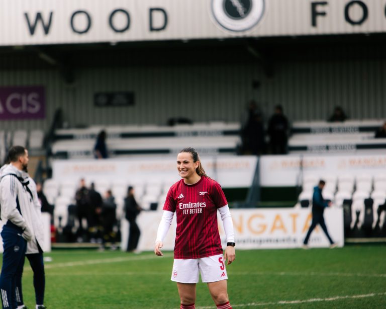 Arsenal defender Laia Codina smiles during the pre-match warm-up, standing on the pitch in Arsenal's red training shirt over a white base layer and white shorts. The blurred stands and stadium signage form the background, with staff and players visible around the edge of the pitch.