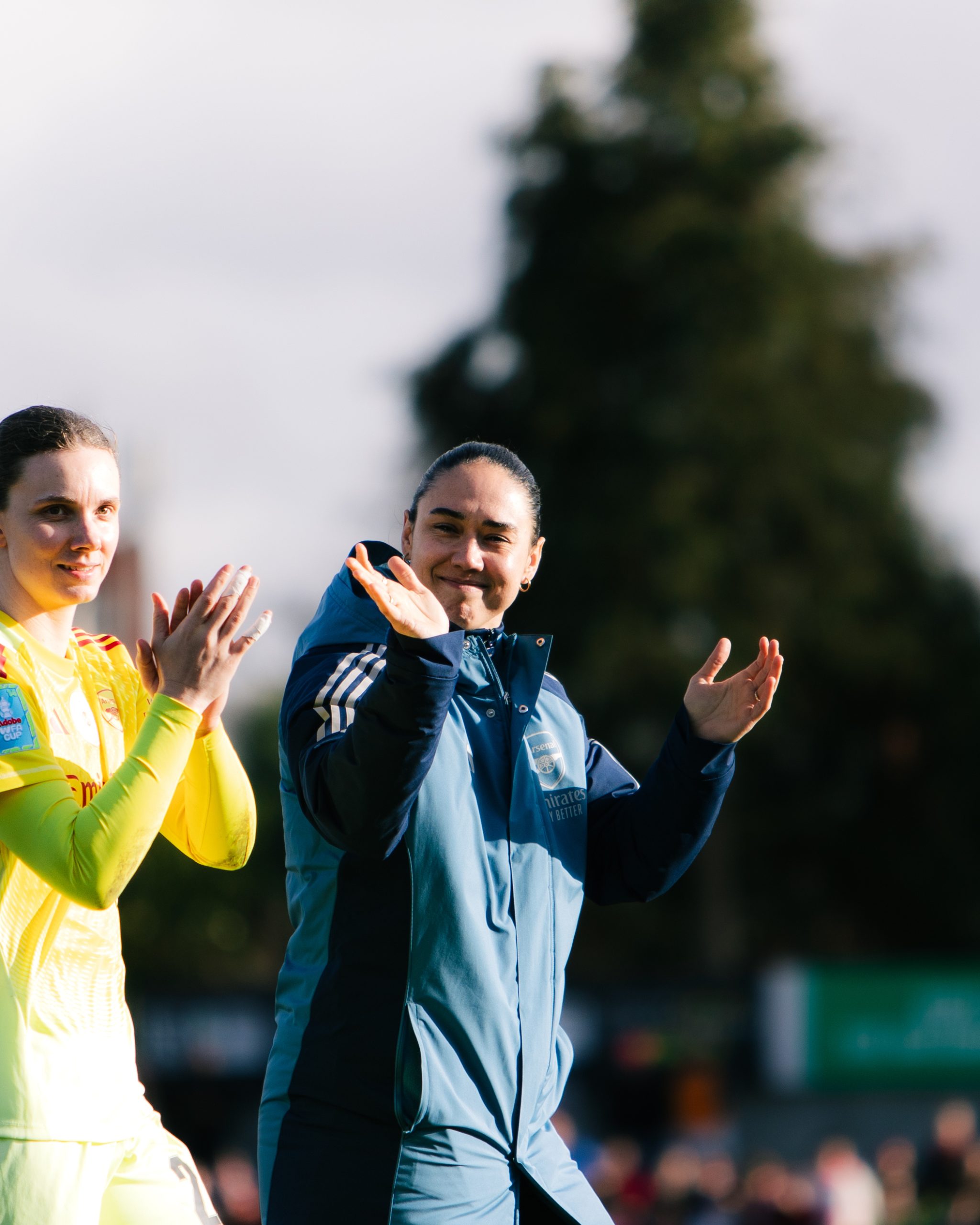 Manuela Zinsberger, wearing an Arsenal training jacket, applauds supporters with a smile after a match while standing alongside teammate Anneke Borbe in a yellow goalkeeper kit. Both players are clapping as they acknowledge the crowd, with trees and blurred spectators in the background.