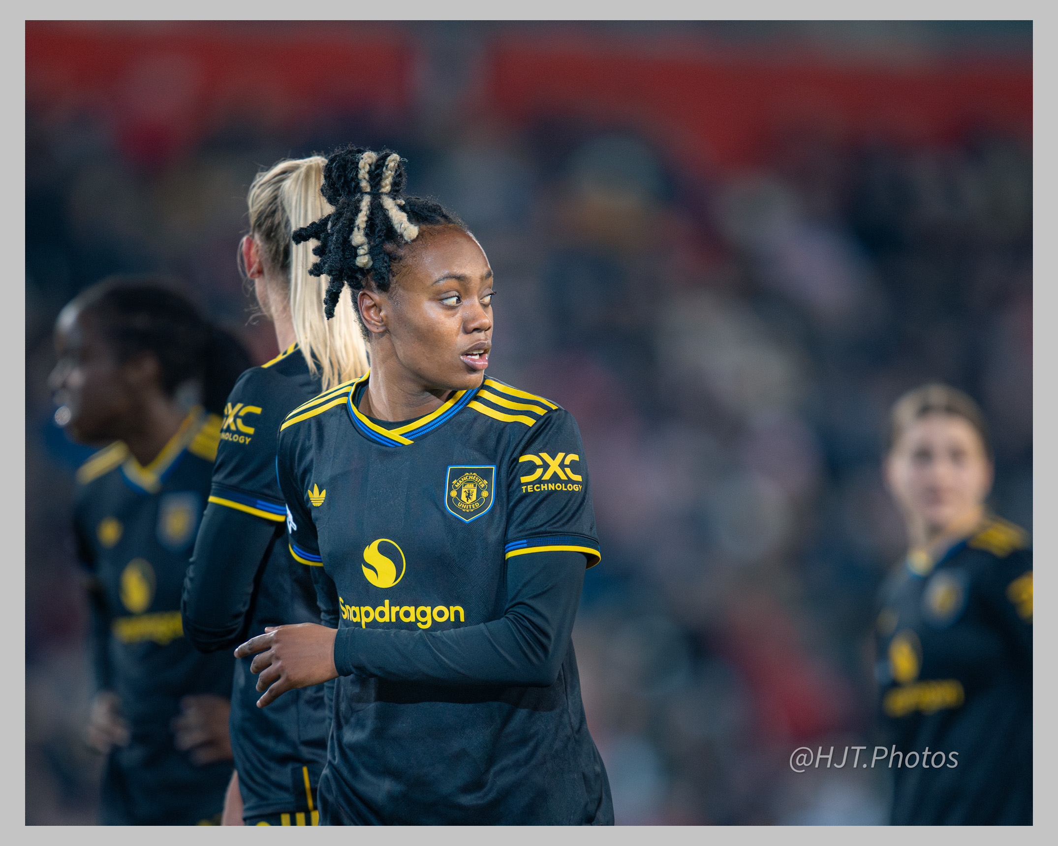 Melvine Malard in Manchester United's black away kit with yellow trim looks across the pitch during a match. She stands in sharp focus in the foreground with her hair tied in a high bun featuring blonde highlights, while three teammates appear blurred in the background against an out-of-focus stadium crowd.