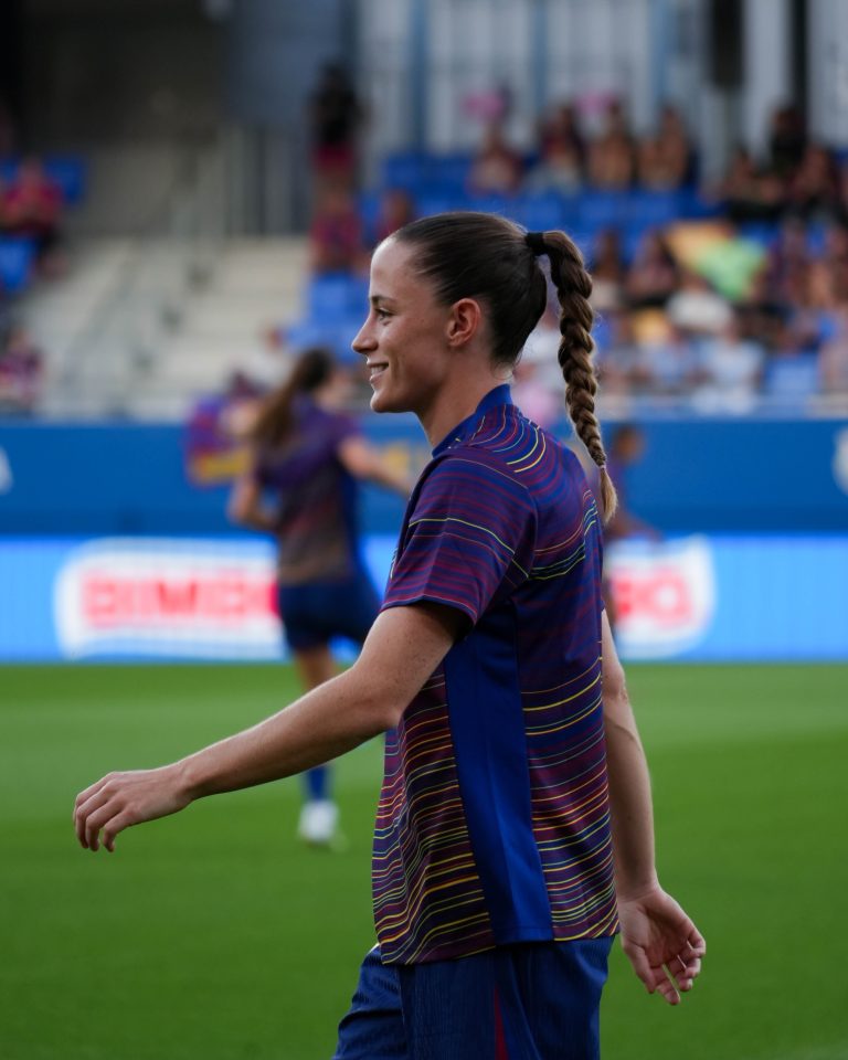 Side-profile photograph of Ona Batlle smiling as she walks across the pitch during pre-match warm-ups. She is wearing a navy and burgundy striped training top with matching shorts, with her hair tied back in a long braided ponytail. The green pitch is in the foreground, while blurred teammates, blue stadium seating and spectators create a soft background.