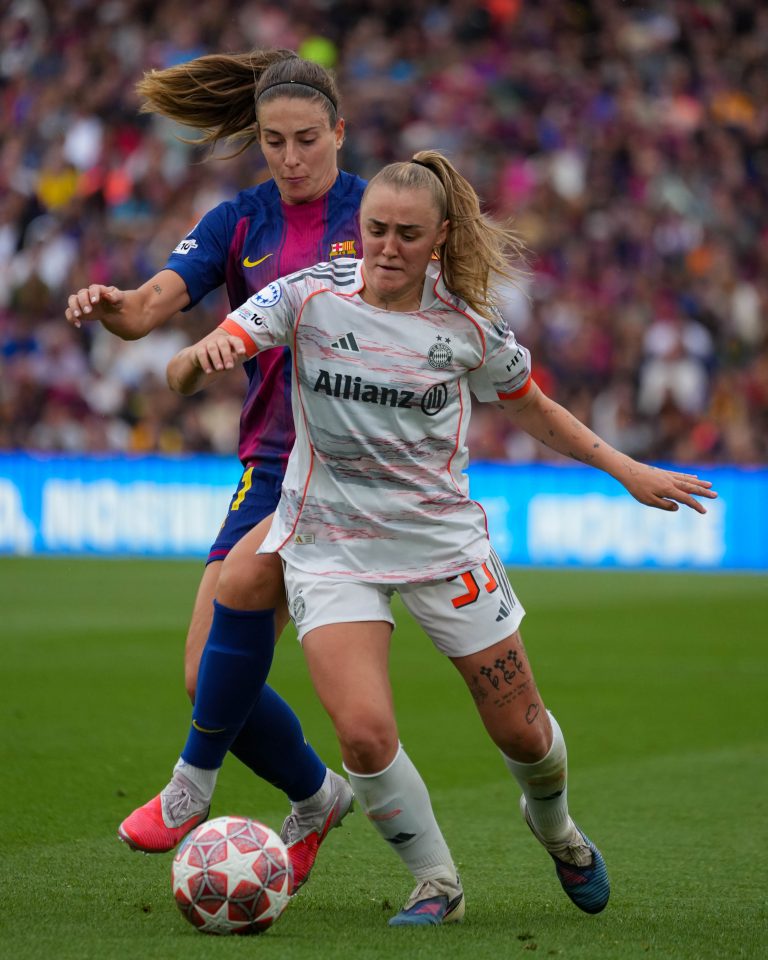 Georgie Stanway as a Bayern Munich player at the time shields the ball from Alexia Putellas, Barcelona player at the time during a UEFA Women's Champions League match. Both players battle for possession, with Stanway using her body to protect the ball as Putellas presses closely from behind in front of a packed crowd.