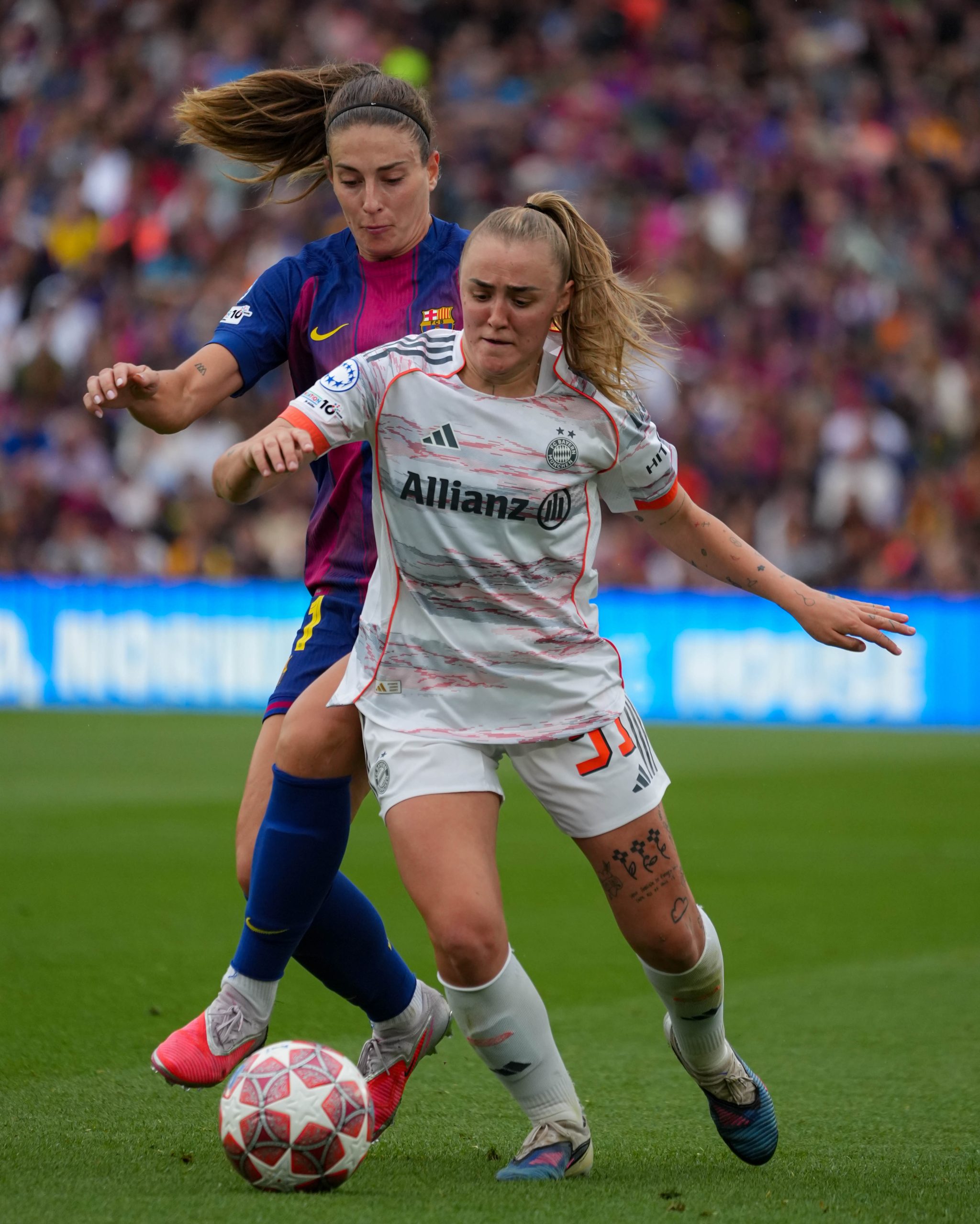 Georgie Stanway as a Bayern Munich player at the time shields the ball from Alexia Putellas, Barcelona player at the time during a UEFA Women's Champions League match. Both players battle for possession, with Stanway using her body to protect the ball as Putellas presses closely from behind in front of a packed crowd.