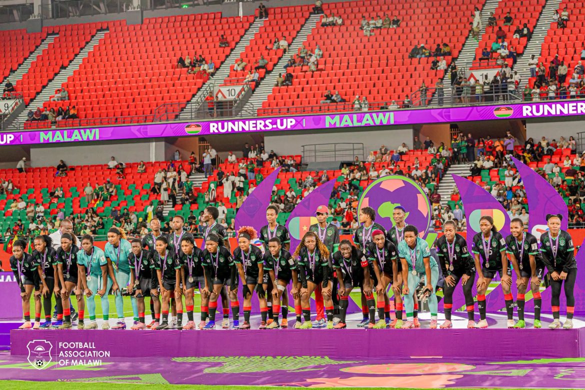 Malawi women’s national team players stand on the runners-up podium wearing medals after the WAFCON final, with supporters and red stadium seats visible behind them.