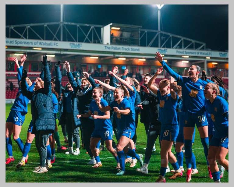 Birmingham City Women players and staff celebrate together on the pitch, raising their arms and cheering in front of the stands at Walsall FC.